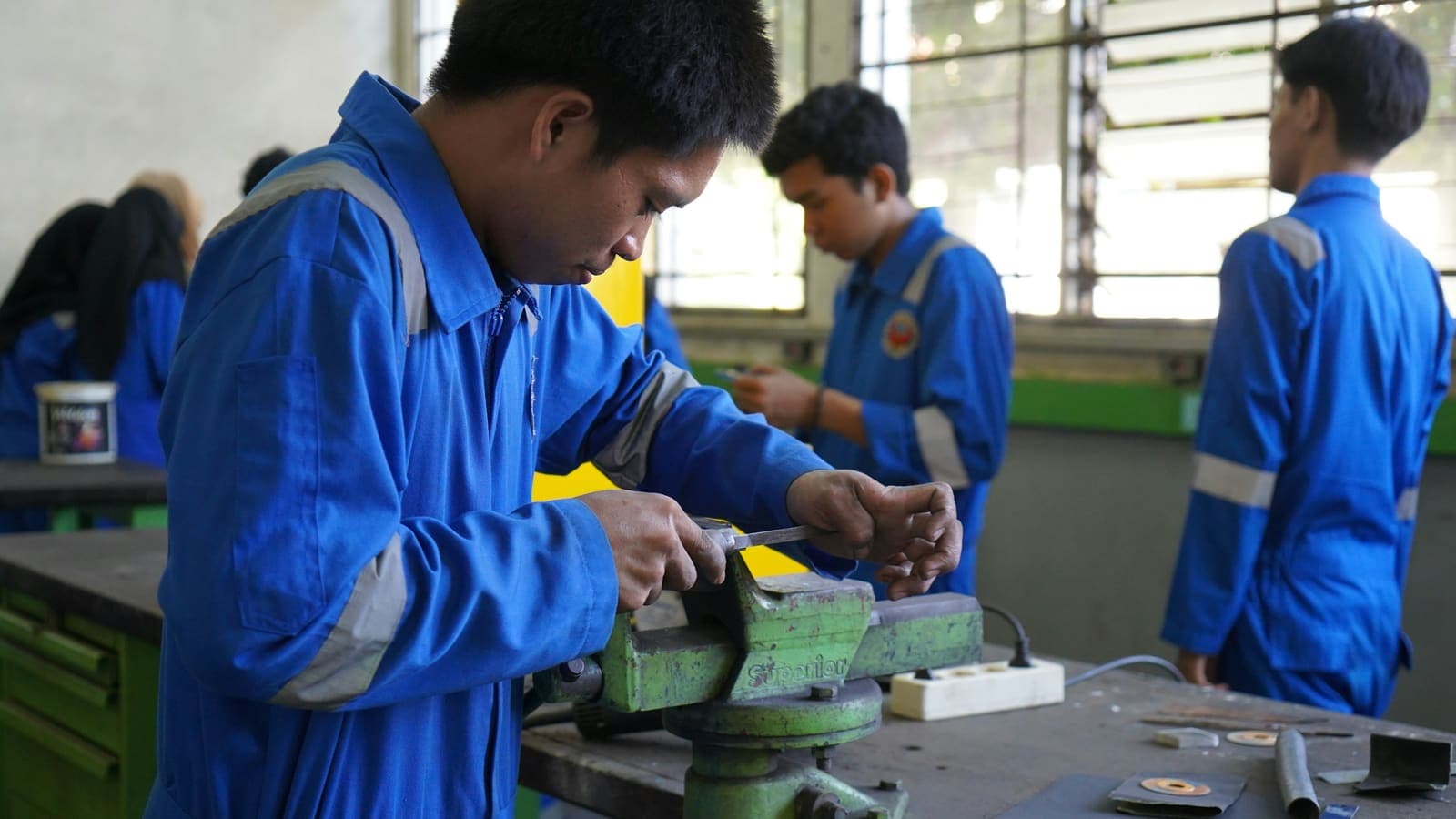 Des apprentis en combinaison travaillent une pièce à l'étau dans un atelier de formation.